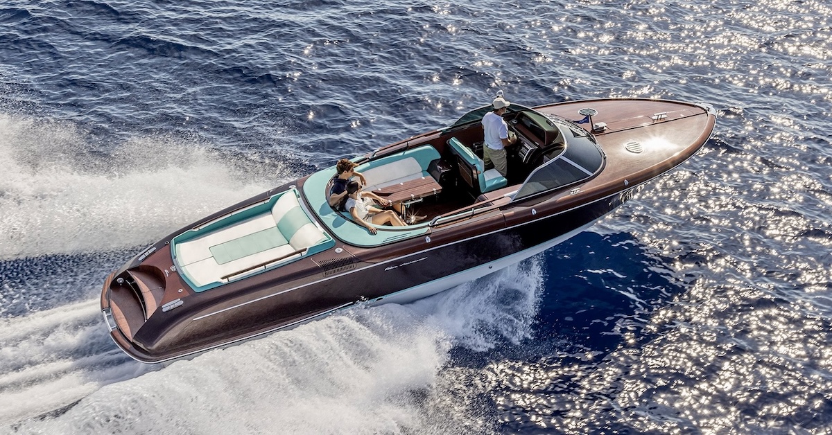 An elegant, wooden speedboat travels at considerable speed over sun-speckled waters. The predominant colour of the boat is 'mahogany' with teal-green trim. Two people are seated in the aft passenger compartment. The person driving the boat is located forward of them, on the left side of the boat where the steering wheel is located. The boat is moving from left to right in the frame and is seemingly photographed from an elevated position, as would be the case if it were from a helicopter. (📸 Riva Yacht)