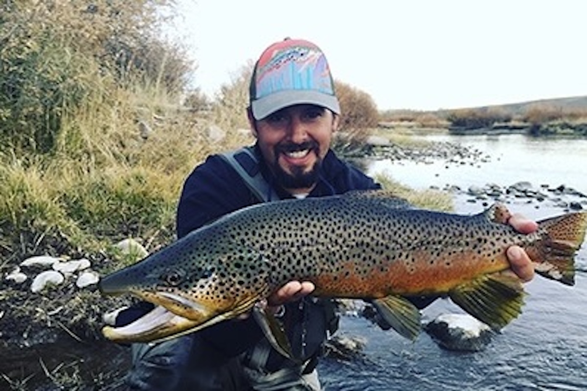A smiling man holding a northern pike he has just caught is pictured in front of the river from which the fish was presumably taken.