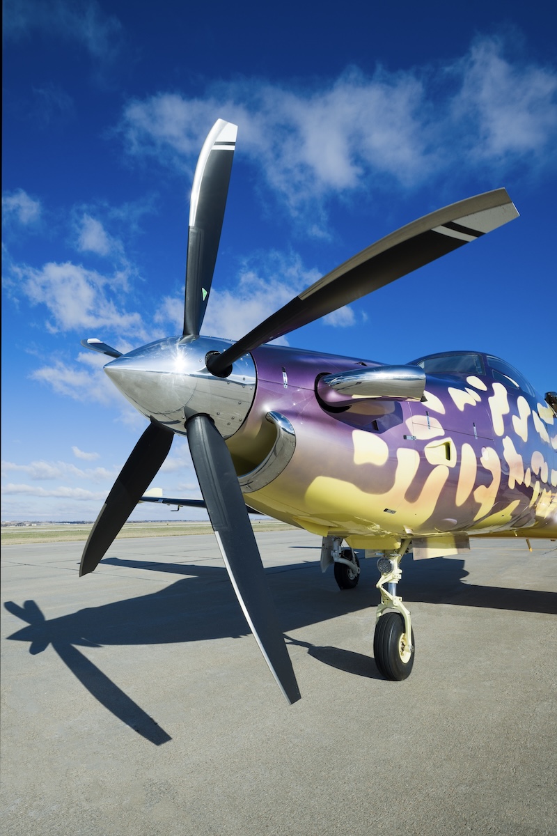The nose of a Pilatus PC-12 turboprop which is parked on an airport ramp. It features a disproportionately large, five-blade black propeller.