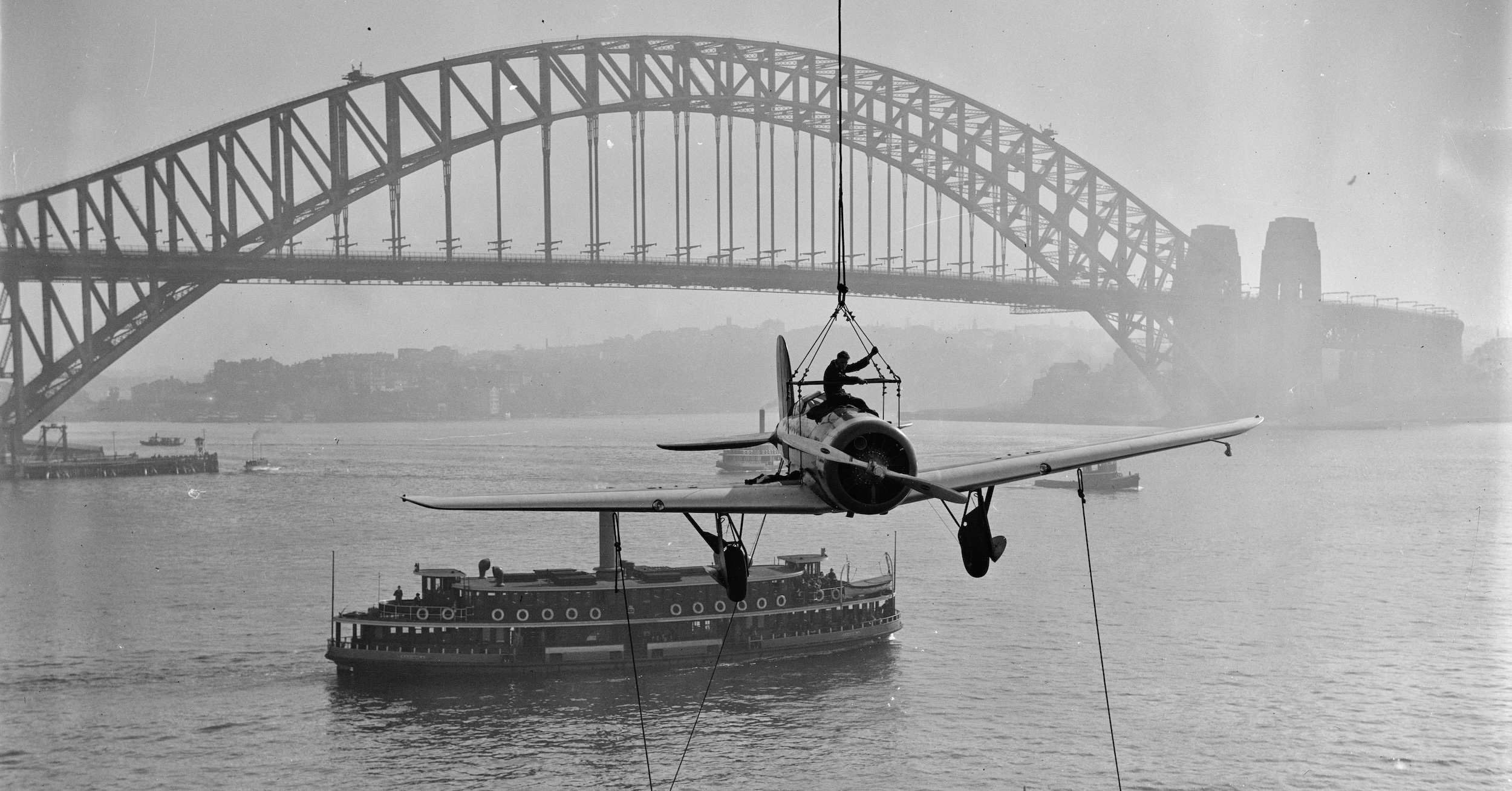 A vintage, low-wing monoplane aircraft is suspended from a crane while being unloaded from a ship's deck. The Sydney Harbour Bridge is in the background. (📸 Sam Hood, from original glass negative, Mitchell Library, State Library of New South Wales, ON 204/Box 68/3.)