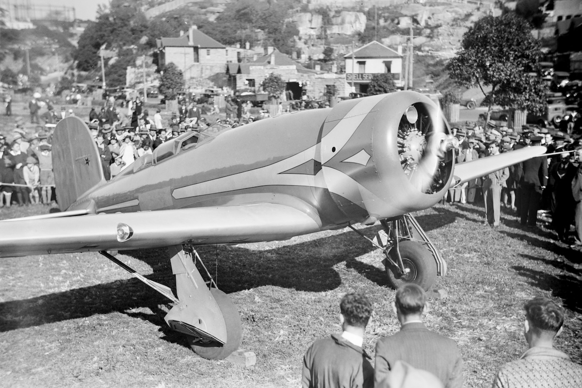 A vintage, low-wing monoplane with its engine running prepares to take off from a grass airstrip. A crowd of mostly men in the background watches.