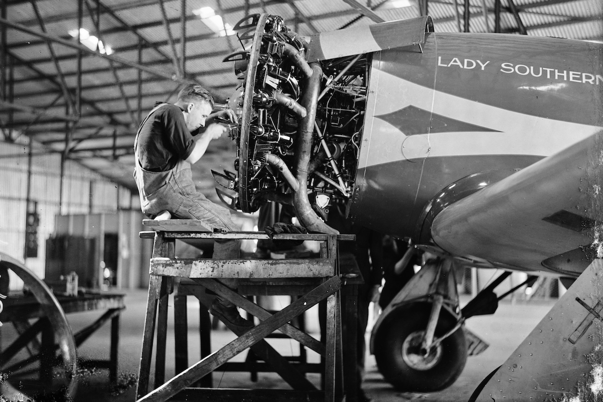 A man works on the uncowled radial engine of a vintage, low-wing monoplane. He is seated on a platform, raising him up to engine height. 