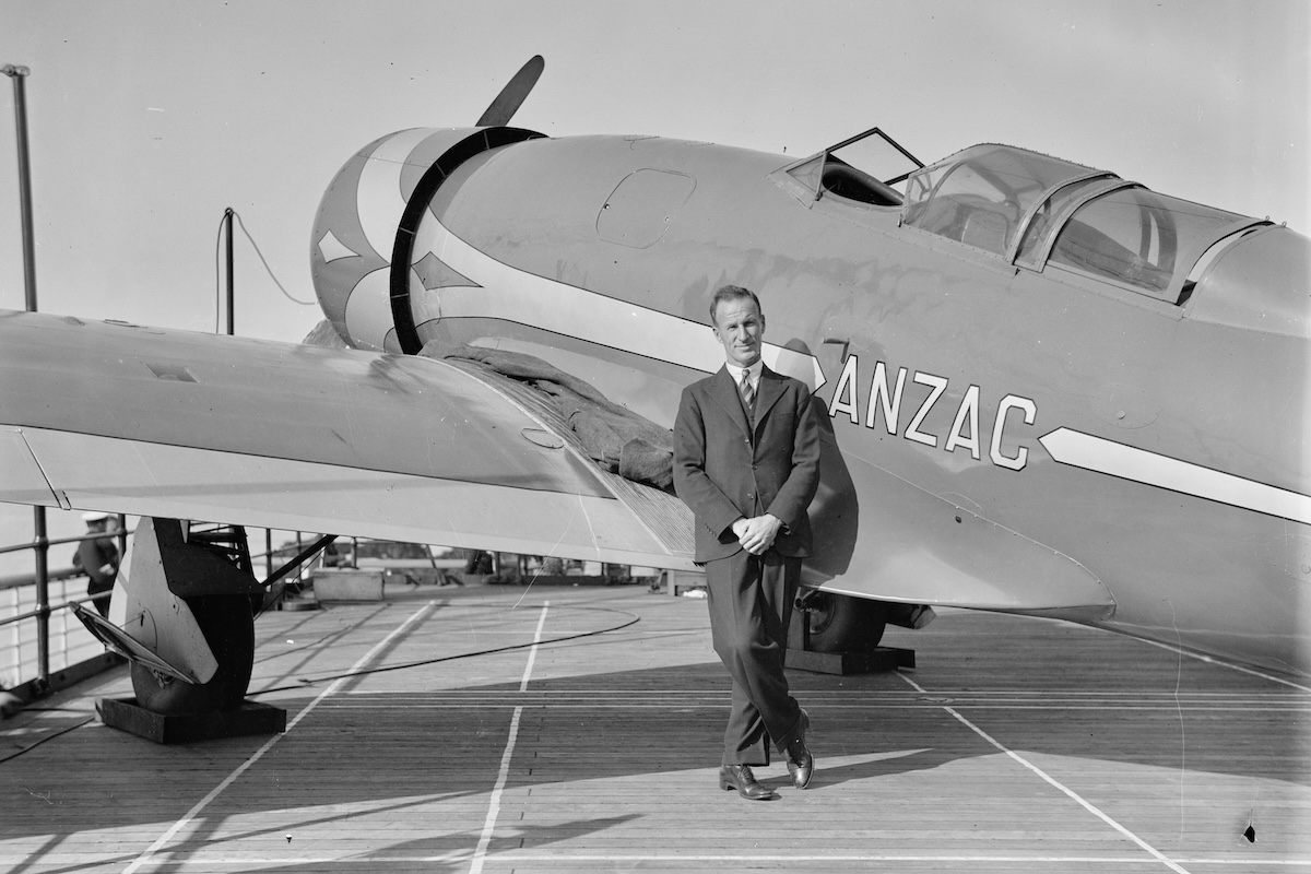 A man in a suit stands in front of a vintage, low-wing monoplane aircraft, aft of the wing. He is looking directly at the camera.