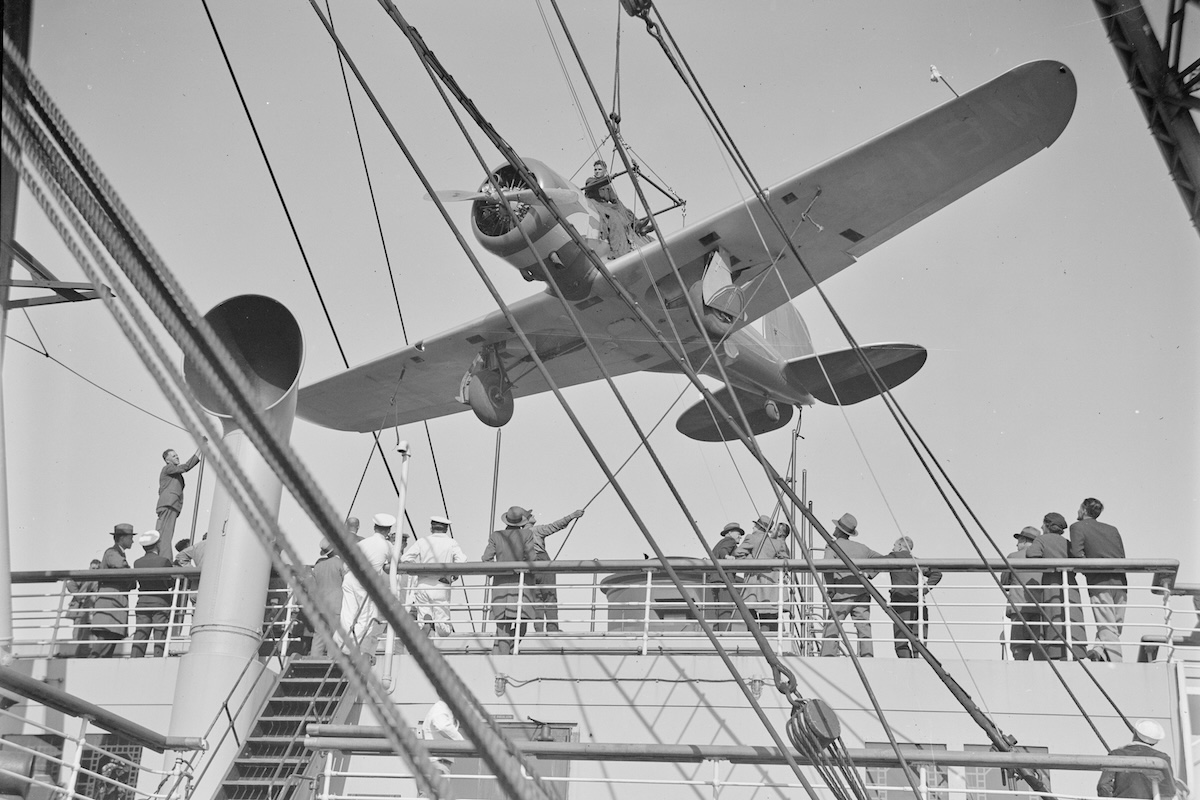 A vintage, low-wing monoplane is suspended from a cable over the deck of a ship as dock workers look on and guide it with ropes.