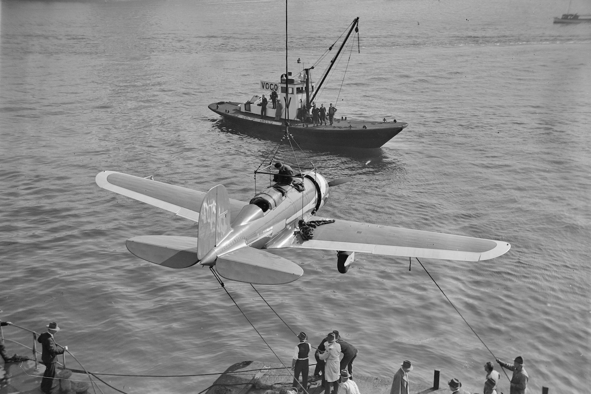 A vintage, low-wing monoplane is suspended from a cable over a body of water. A small ship is visible just beyond the aircraft. 