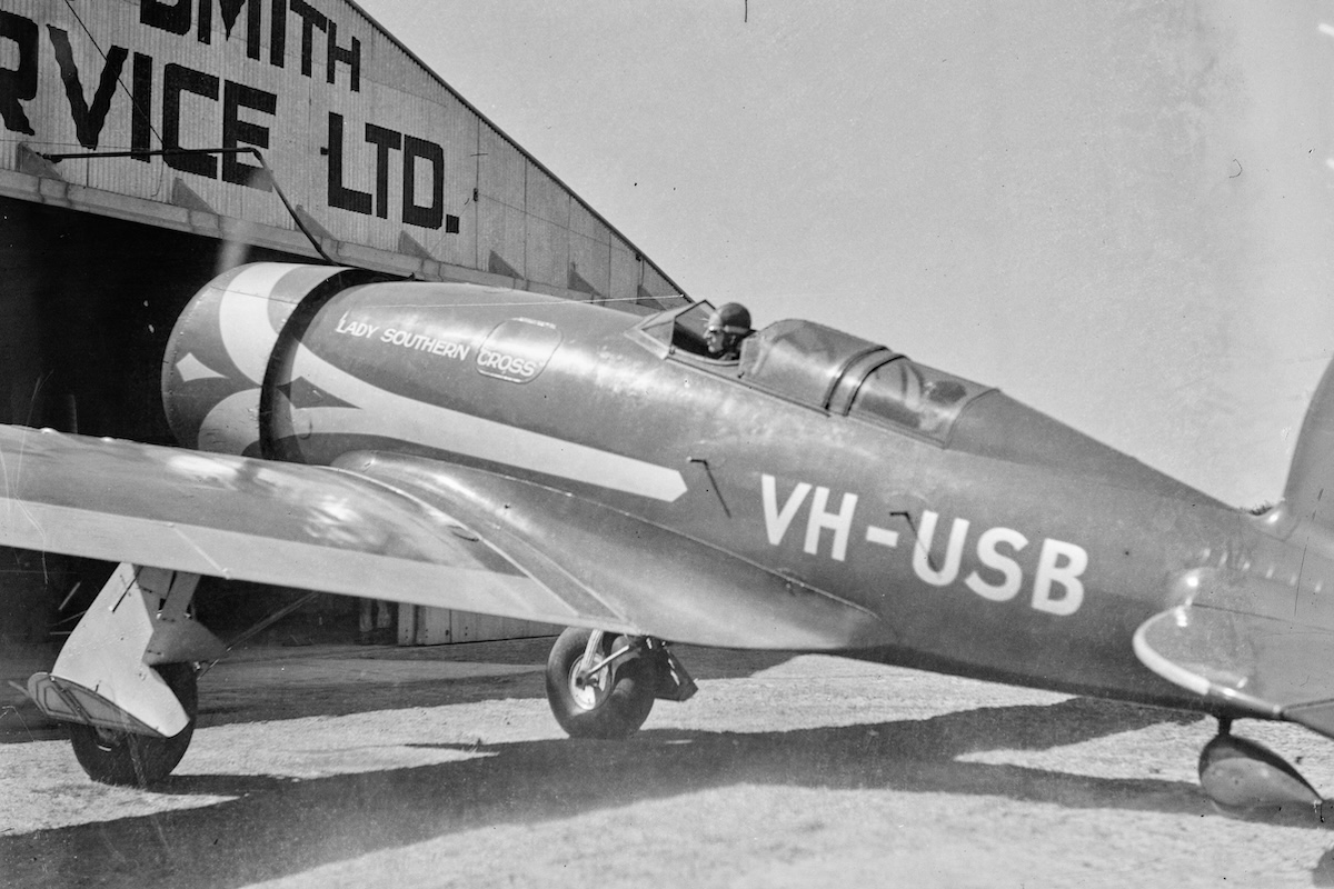 A vintage, low-wing monoplane with its engine running is parked facing the open door of a hangar. The pilot is visible in the cockpit. 