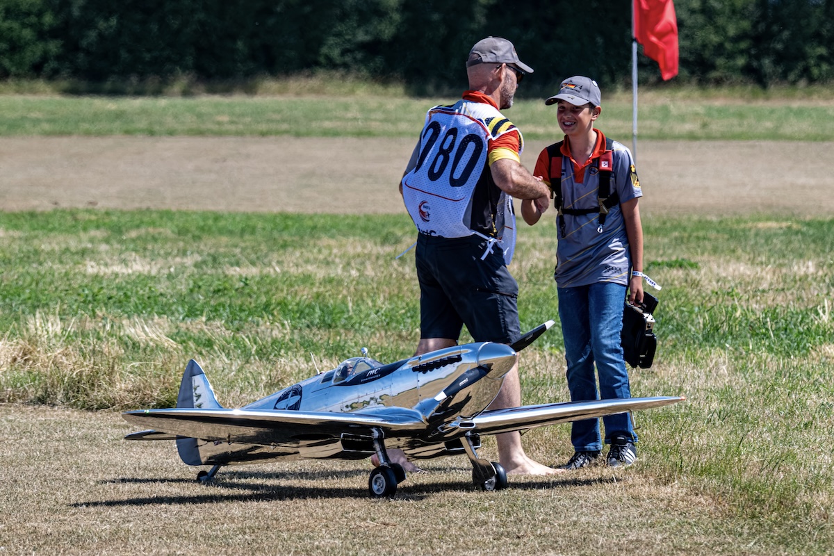 A man shakes the hand of a smiling boy. They are standing beside a model of a Supermarine Spitfire on a grass landing strip. 