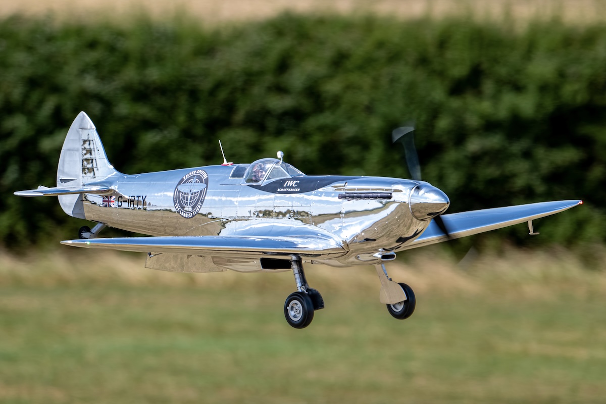 A model of a Supermarine Spitfire, in a chrome-like finished meant to reflect polished aluminum, flies from left to right in the frame. 