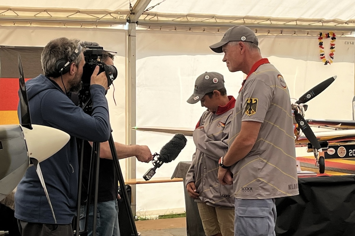 A boy and a man talk with reporters with a microphone who are interviewing them. A model aircraft is behind them, on a table. 