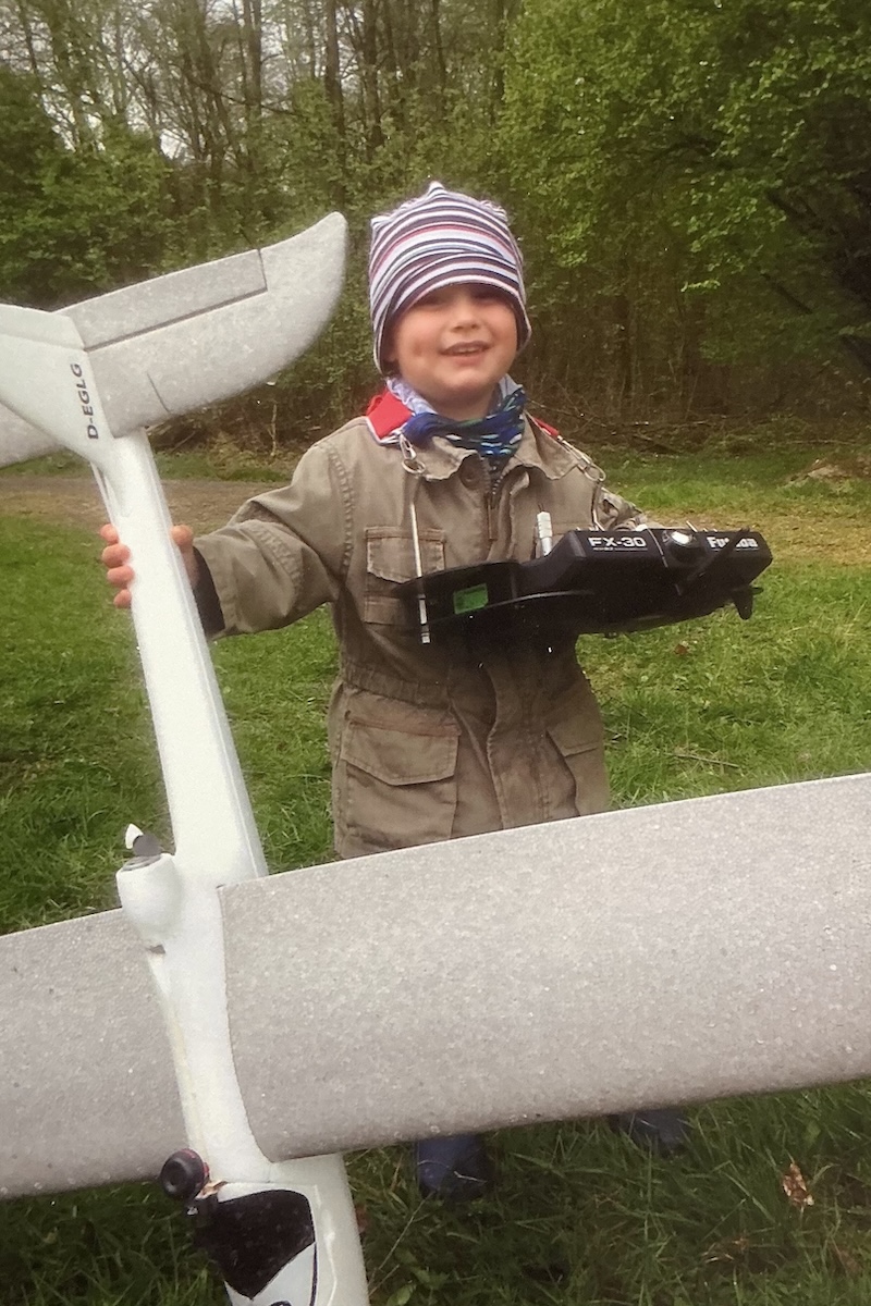 A small boy dressed in winter-appropriate clothing holds a radio-control transmitter while also holding the tail of a foam glider in his right hand.