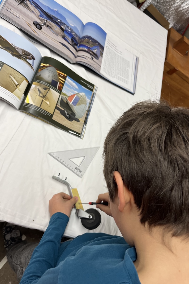 A boy works on a model aircraft tailwheel at a workbench while referring to books as a reference. The bench top is covered with paper. 
