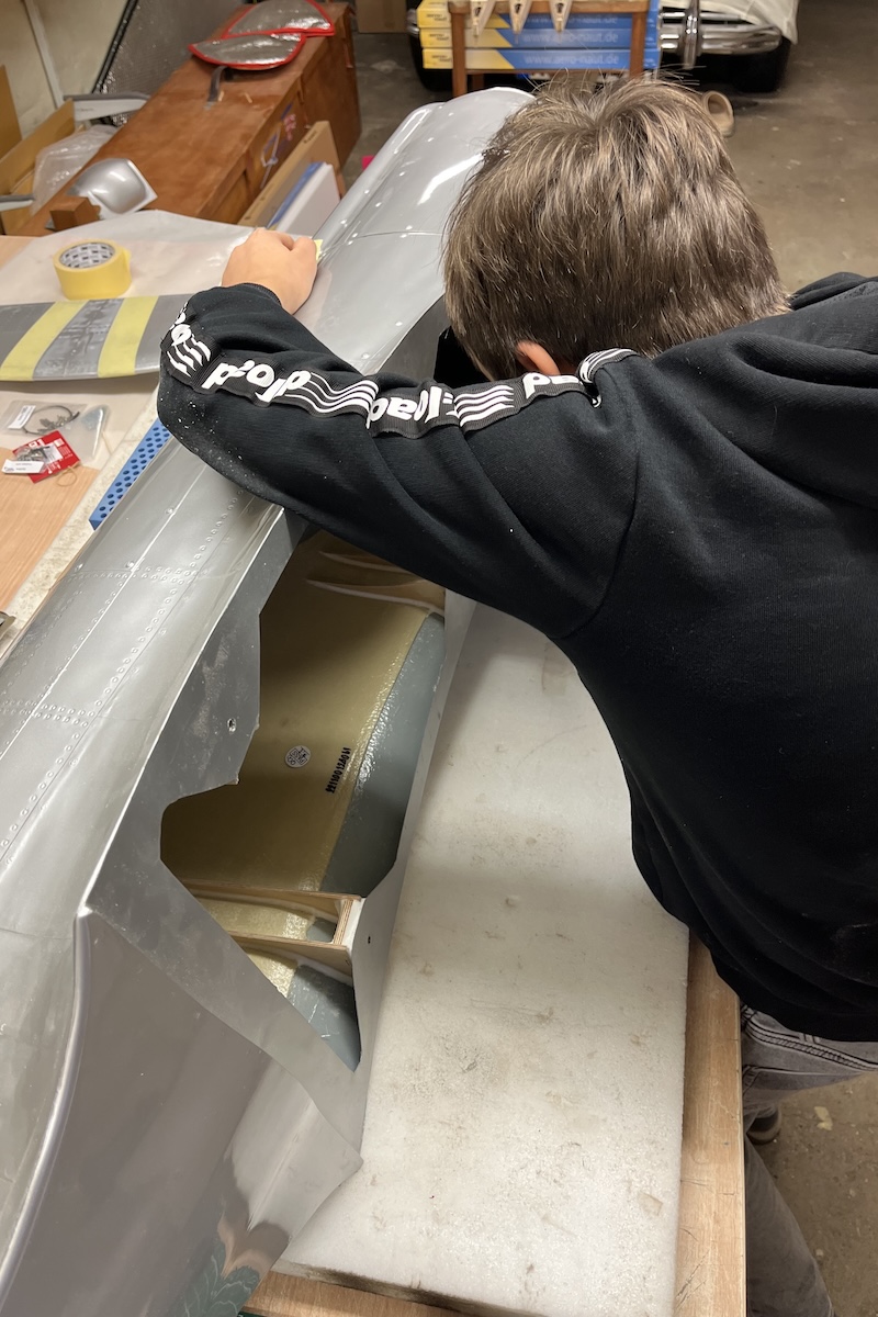 A boy works inside the fuselage of a silver-coloured model aircraft which is on its side, reaching through the underside where the wing normally goes. 