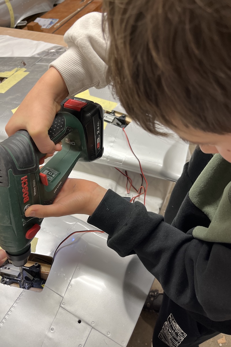 A boy holds a power tool, resembling a drill, while working on what appears to be the underside of a wing of a model aircraft. 