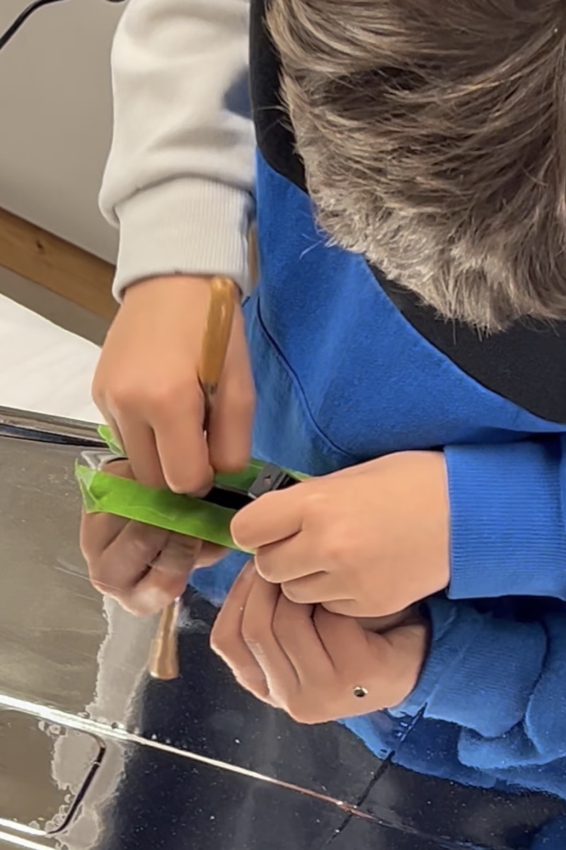 A boy holds a small knife in his right hand while working on the shiny surface of what appers to be a model aircraft wing. 