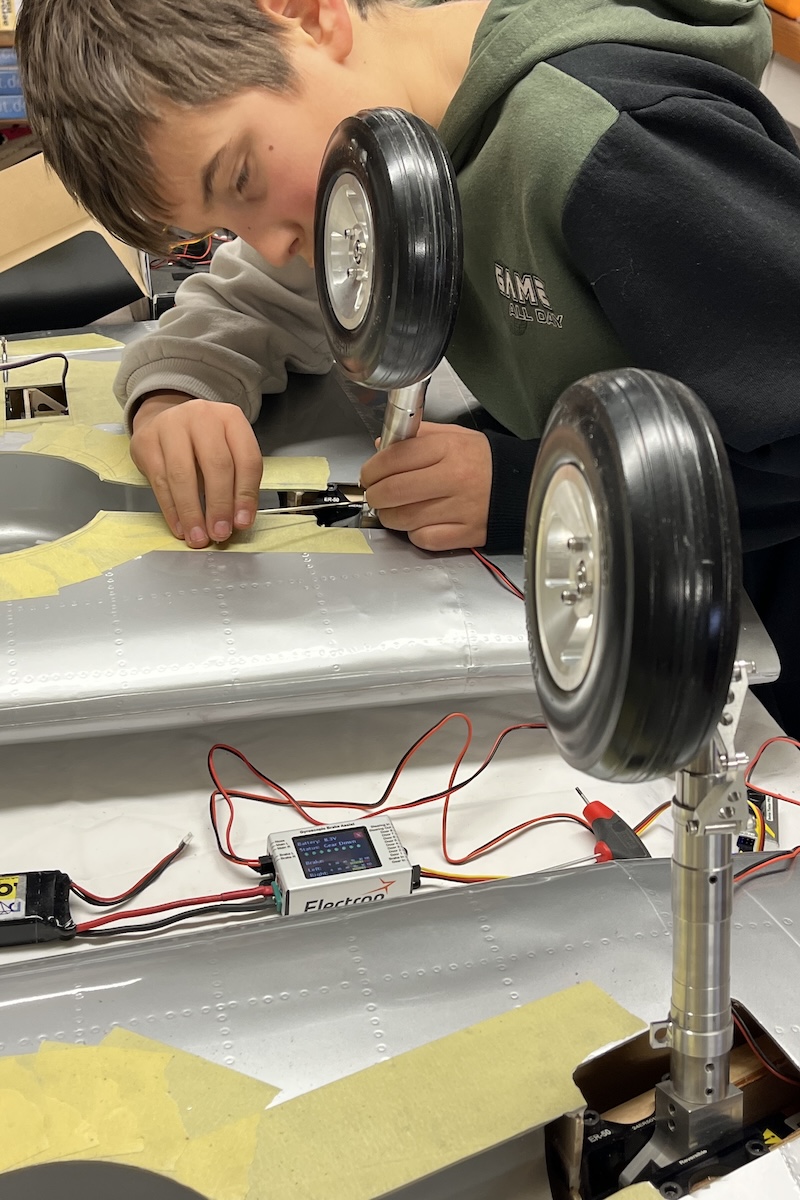 Using a small screwdriver, a boy works on the extended, retractable landing gear extending upwards from two model aircraft wing halves positioned on a workbench. 
