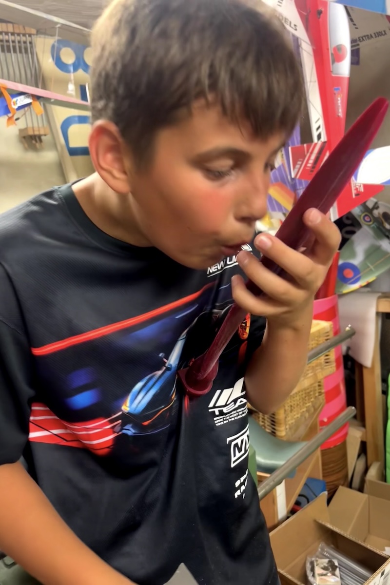 A boy blows on, as if to dry it or remove dust, the blade of a reddish-coloured, 3D-printed propellor blade from a scale model aircraft. 