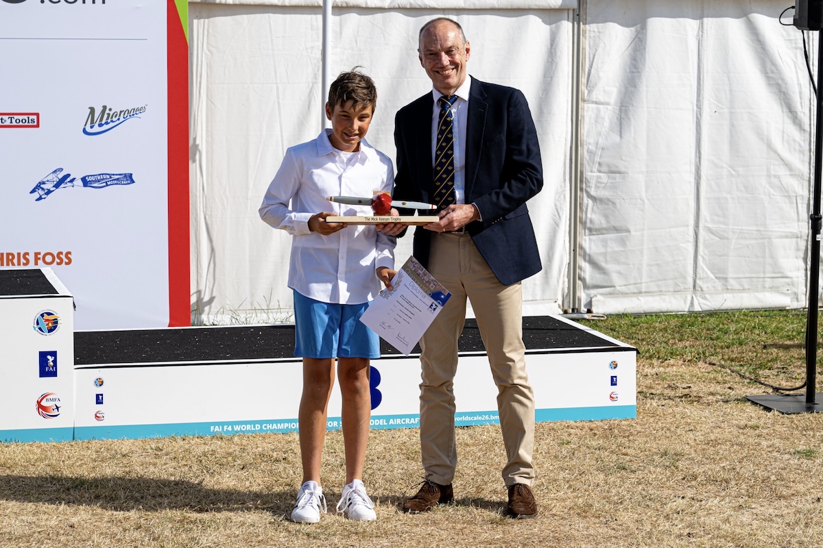 A man, on the right, presents a trophy and a certificate to a boy, on the left. It's a sunny day and both are smiling.