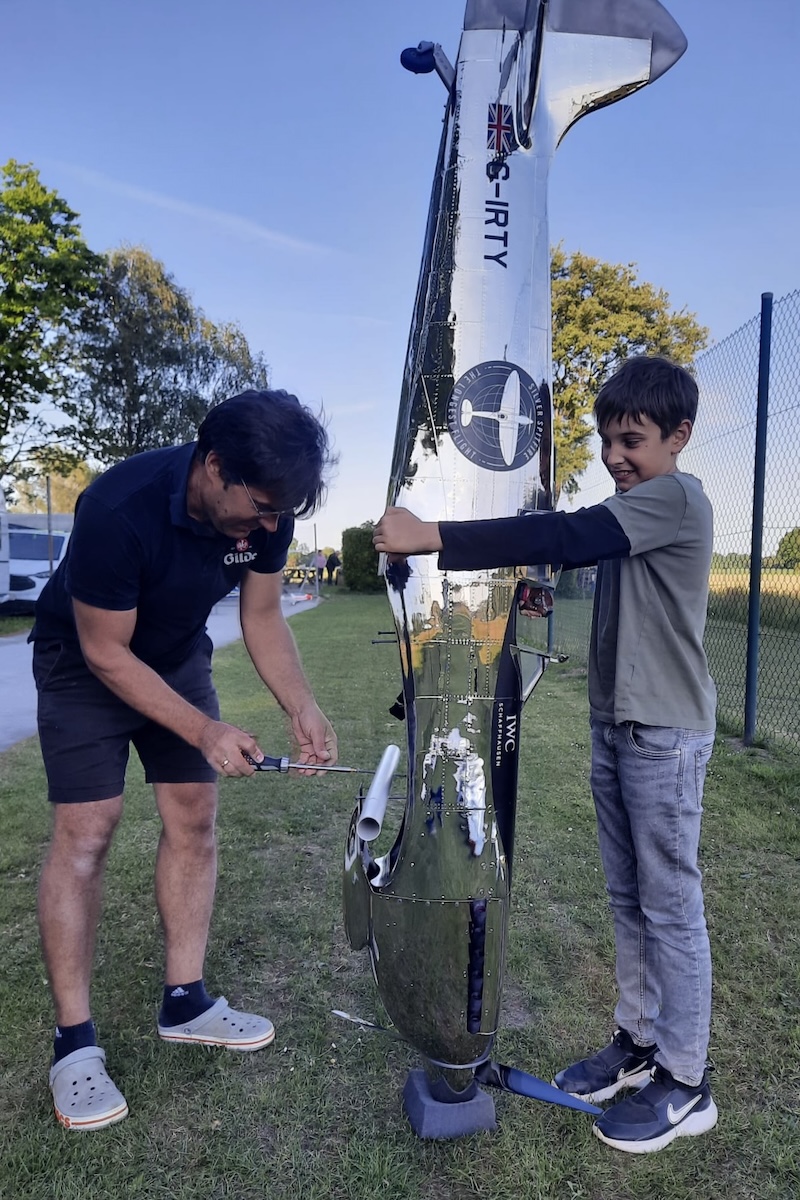 A boy holds the fuselage of a model aircraft, with its nose pointed to the ground, while a man attaches an aluminum tube where the wing usually goes. 