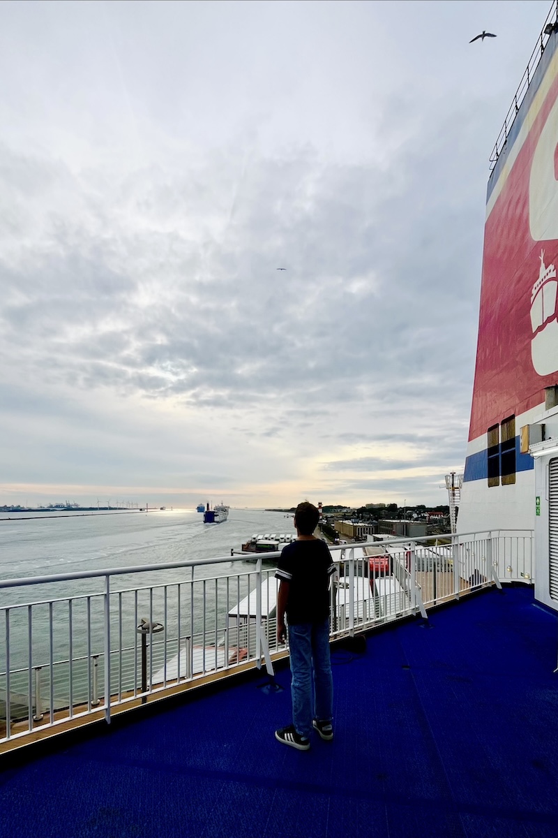 A boy stands next to the funnel of a ferry, appearing to be preparing for departure. He is looking at another ship that is departing. 