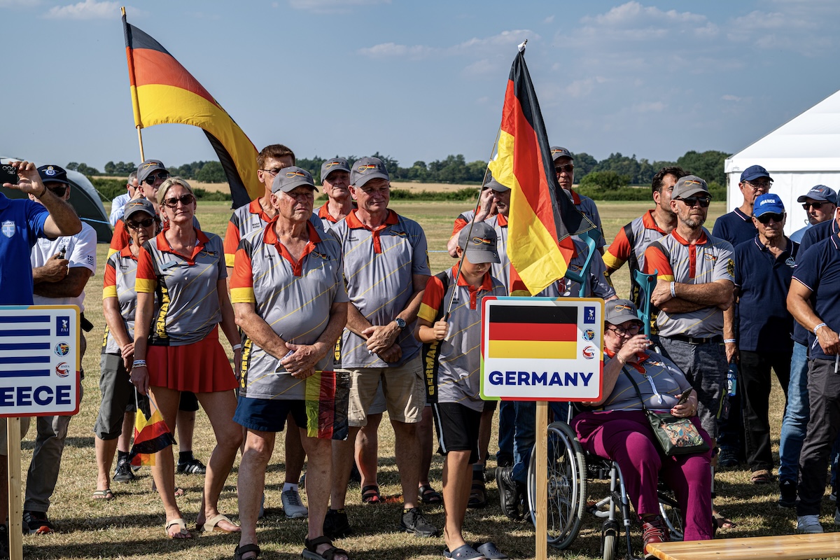 A group of people holding German flags stand behind a sign that says Germany along with the German flag. They are all wearing team jerseys.
