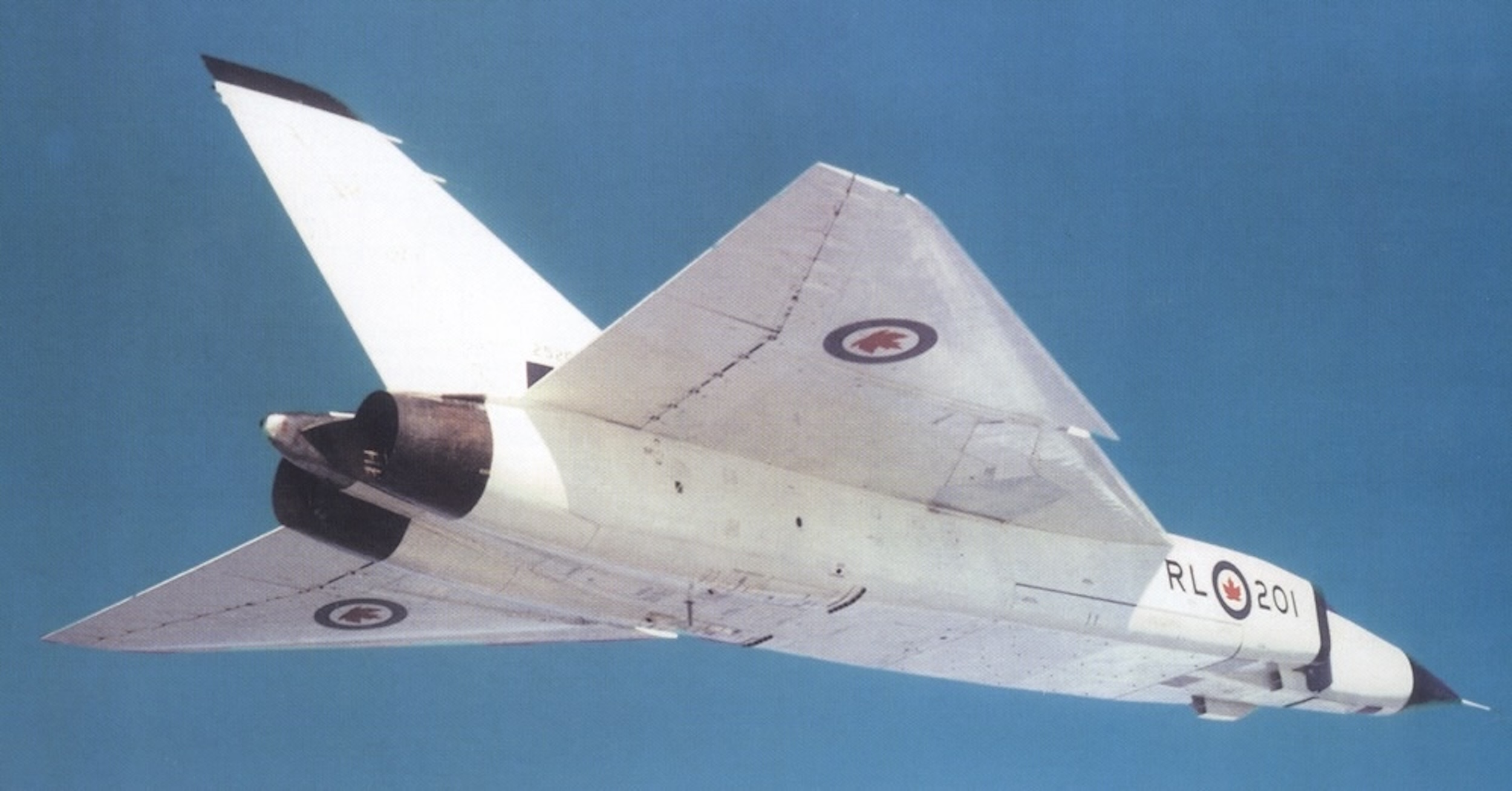 A white, twin-engine, delta-wing jet flies in a gin-clear, blue sky. The photograph is taken from just below, just aft, and just to the right of the aircraft, exposing the right side of its fuselage. Royal Canadian Air Force roundels are painted on the lower surface of both wings and on the side of the fuselage just below where the cockpit is located. On either side of the roundel on the fuselage are the letters « RL » and « 201 ». Caption from source: « The first of six Arrows produced on the near-production-ready Avro Canada assembly line in Malton, Ontario, in the late 1950s. (📸 Avro Canada) »