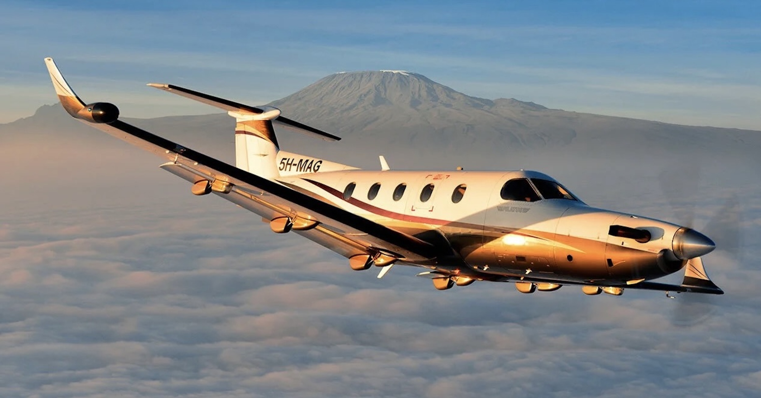 An in-flight photo of a Pilatus PC-12 as it banks away from the camera position. Mount Kilimanjaro, with only a dusting of snow on its peak, is visible in the background. The lighting is such that it looks like the photo was taken either at sunrise or sunset. The aircraft is mostly white, with a curvaceous, graphic pattern consisting of various shades of gold on the lower part of the fuselage and the tip of the vertical stabiliser. The registration « 5H-MAG » is painted on in the vertical stabiliser in black, bold letters. There is a cloud deck below the aircraft and a mostly blue sky with some high, streaky clouds present. Original caption from source: « The Coastal PC-12 with Mount Kilimanjaro in the background. » (📸 ©2017 Jon Davison | Eye in the Sky Productions) (📸 ©2017 Jon Davison | Eye in the Sky Productions)