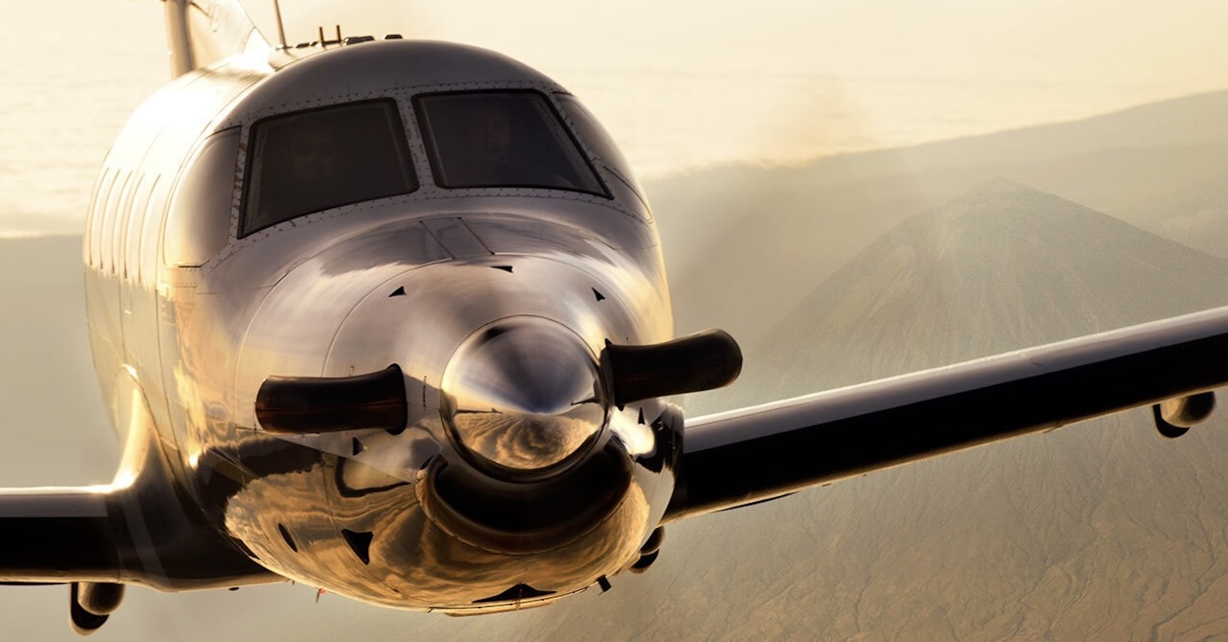 An in-flight photo of a Pilatus PC-12 as it is almost directly in the camera position. The tightness of the crop — the fuselage almost entirely fills the frame — obscures most details of the aircraft in terms of colour scheme and registration. Faint outlines of rugged mountains are visible in the distance. The sky appears somewhat hazy, and the light is that of sunrise or sunset. Original caption from source: « The business end of the Pilatus PC-12 photographed near Mount Kilimanjaro. » (📸 ©2017 Jon Davison | Eye in the Sky Productions) (📸 ©2017 Jon Davison | Eye in the Sky Productions)