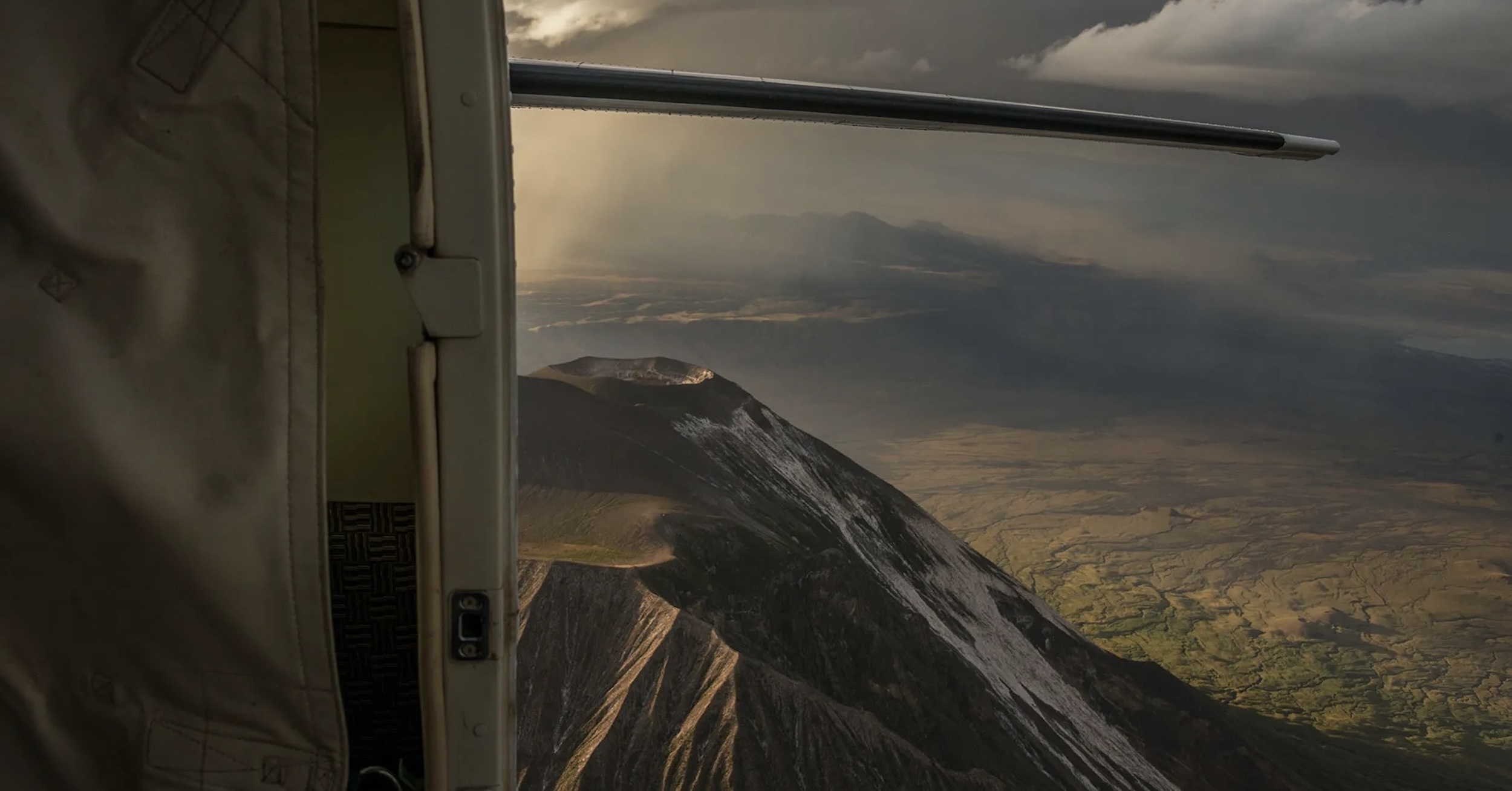 The view from the open cargo door of a Cessna Caravan, looking aft at the crater of Mount Ol’Doinyo Lengai in northern Tanzania. The horizontal stabiliser is visible, as is some snow on the mountain. Beyond the mountain there is green terrain and what could be a lake. Hazy clouds obscure the sky for the most part. Original caption from source: « My view as we cross over the Mount Ol’Doinyo Lengai crater. » (📸 ©2017 Jon Davison | Eye in the Sky Productions)