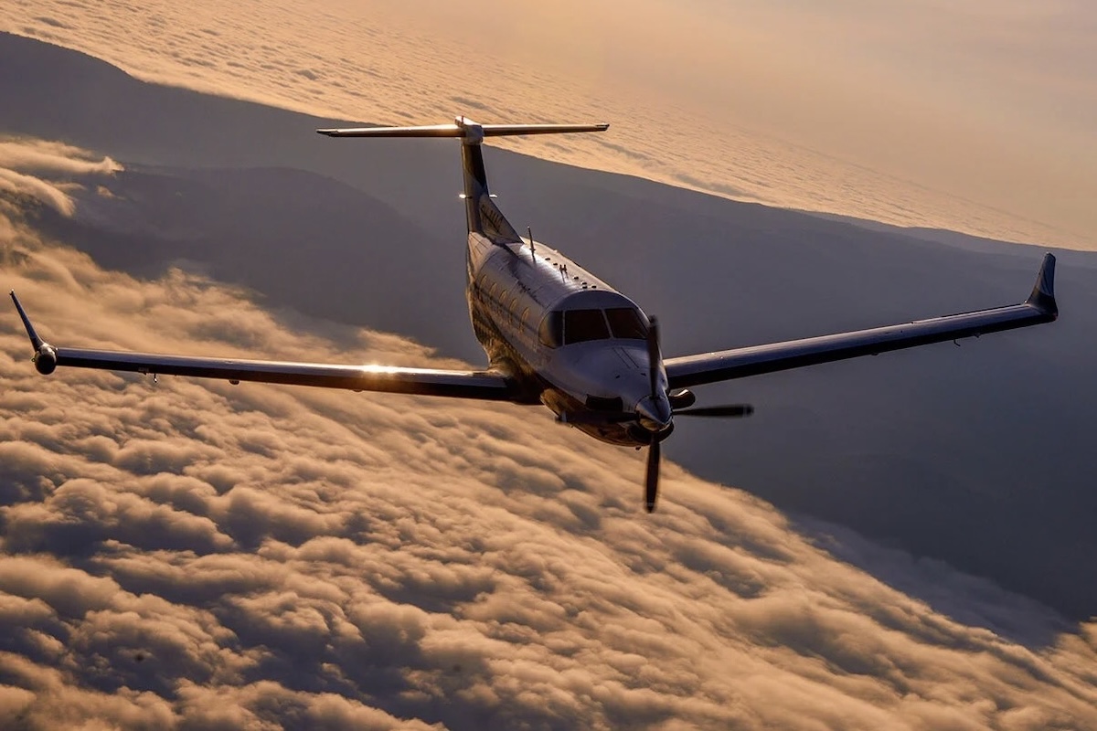 The view from the open cargo door of a Cessna Caravan, looking aft at a Pilatus PC-12 which is flying in formation over a nearly solid cloud deck. The aircraft appears to be banking slightly to the right. Part of the crater of Mount Ol’Doinyo Lengai in northern Tanzania is visible in the background. Original caption from source: « Pilatus PC-12 at sunset under African skies. » (📸 ©2017 Jon Davison | Eye in the Sky Productions)