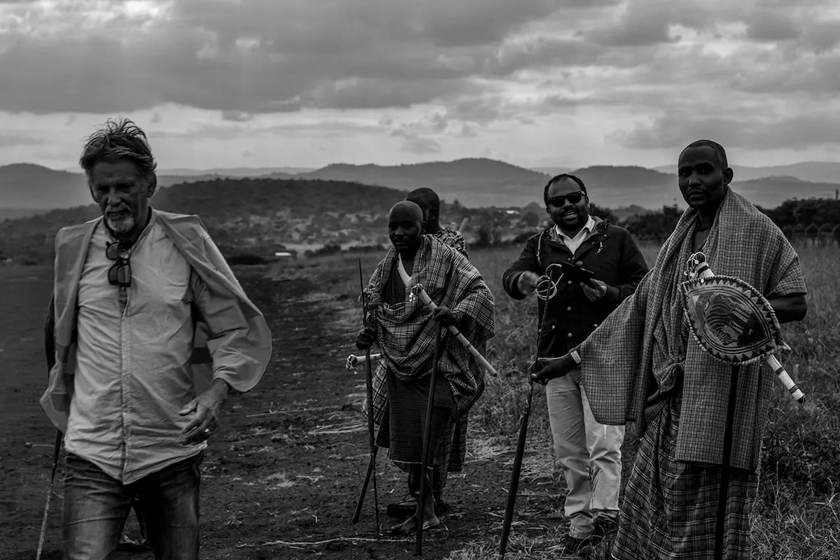 A black-and-white photo of a group of people walking on a scrubby plain. On the left is Jon Davison, who is walking toward and from the right to left in the frame. Slightly behind him, on the right side of his frame and to Jon's left, a group of four people walks in the same direction. Three of the four are Maasai tribesmen wearing their traditional shúkà, whereas the fourth is Mark Mutiso, one of the photo shoot crew members, with a camera and other equipment around his neck.  In the middle distance, a series of rolling hills is visible. Above, the sky seems to be gathering storm, although it's difficult to discern given the lack of colour in the photo. (📸 ©2017 Jon Davison | Eye in the Sky Productions) (📸 ©2017 Jon Davison | Eye in the Sky Productions)