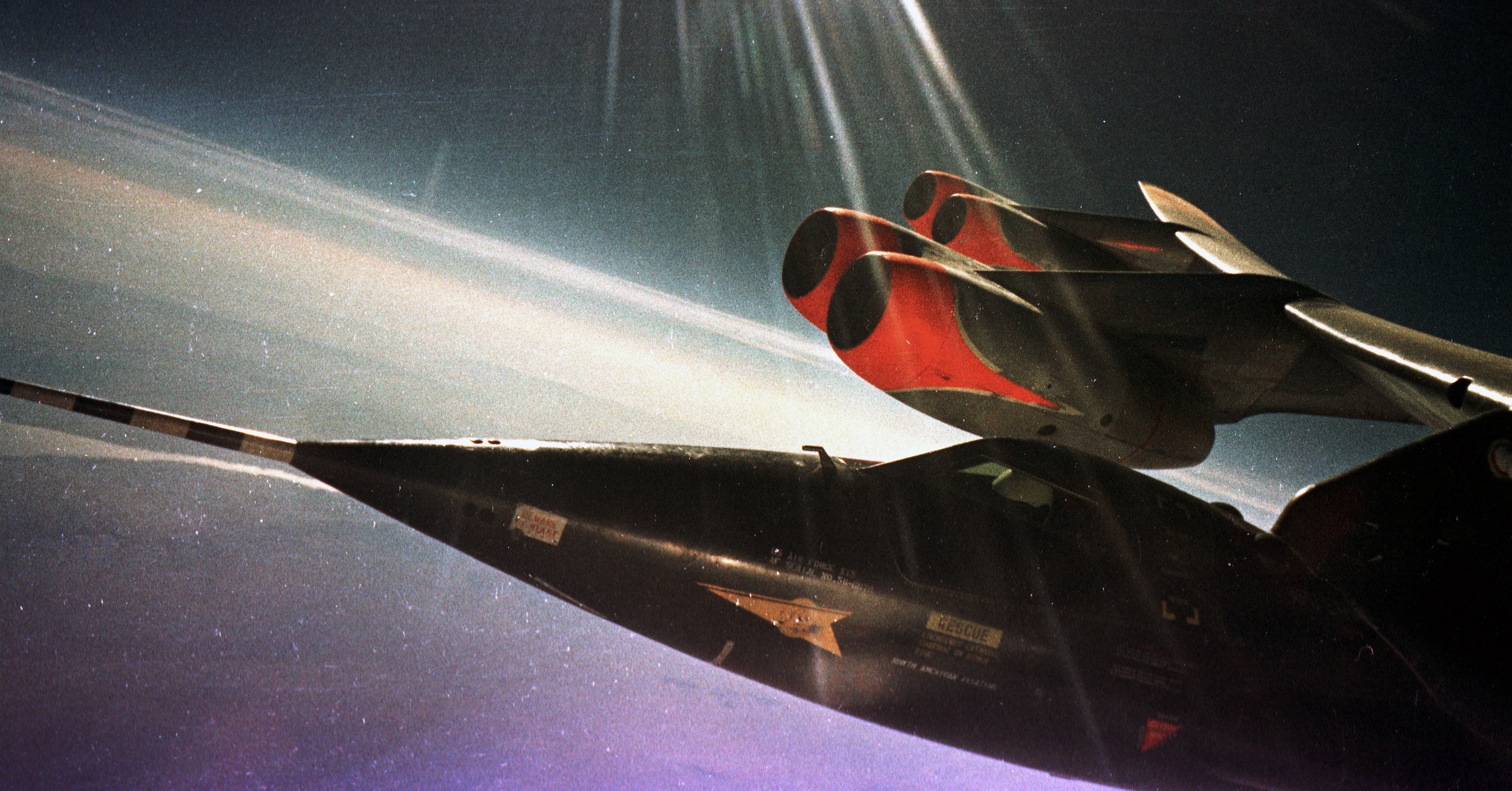 An image of the X-15 experimental space plane suspended from the right wing of the B-52 launch vehicle. Only the nose portion of the aircraft is visible. Close examination of the cockpit area reveals the white helmet of the pilot. The only part of the B-52 visible is the right wing, with its gentle curve upwards under load, and two pods of two engines each. Above, streaks of sun are visible. Far below is the surface of the Earth with a gentle curve of its spherical shape. Original caption from source: « Flight 1-A-18 on August 11, 1960 with pilot Robert M. White in the cockpit of the X-15. » (📸 Air Force Flight Test Center History Office) (📸 Air Force Flight Test Center History Office) »