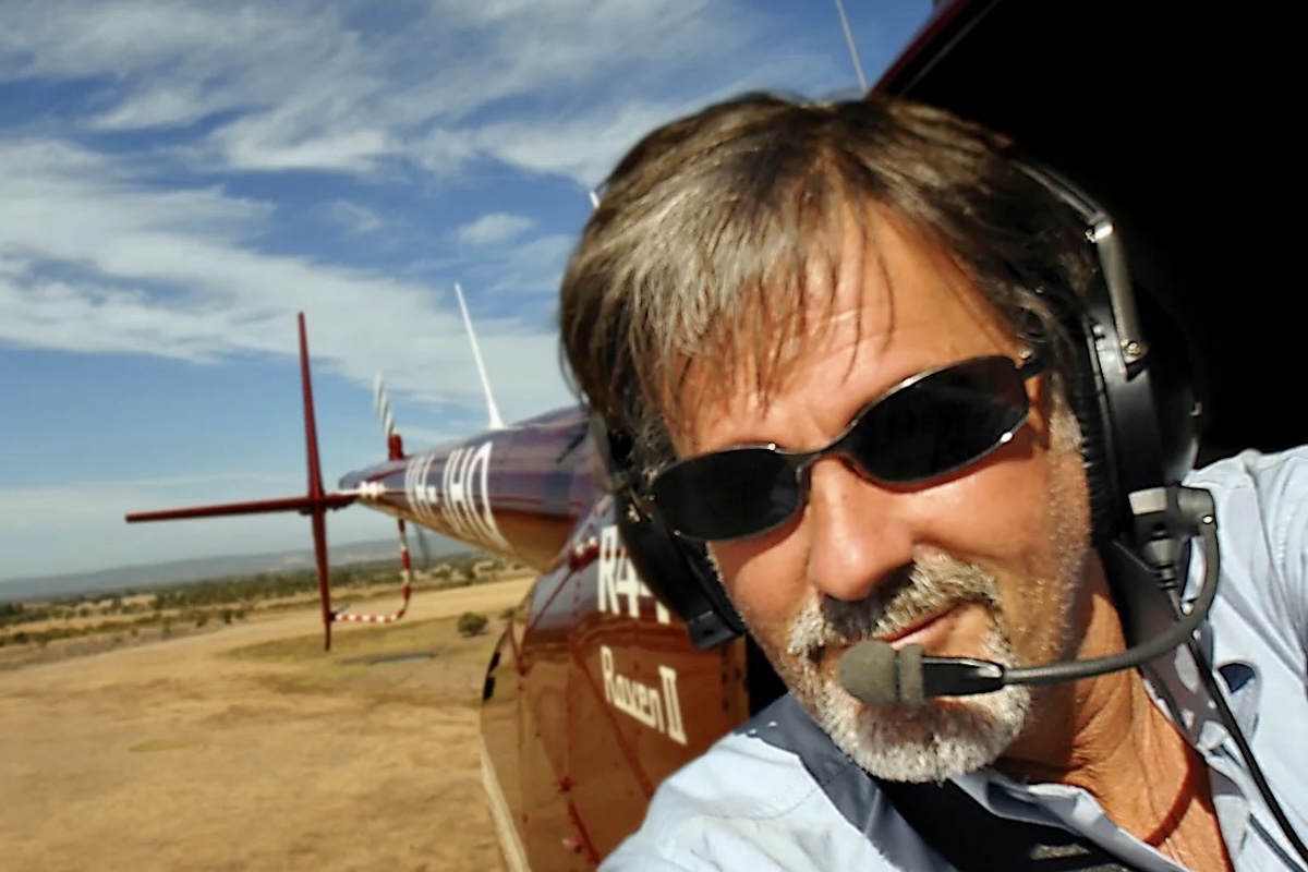 A man with grey hair/beard looks forward and directly at the camera from the open door of a helicopter while it is in flight.
